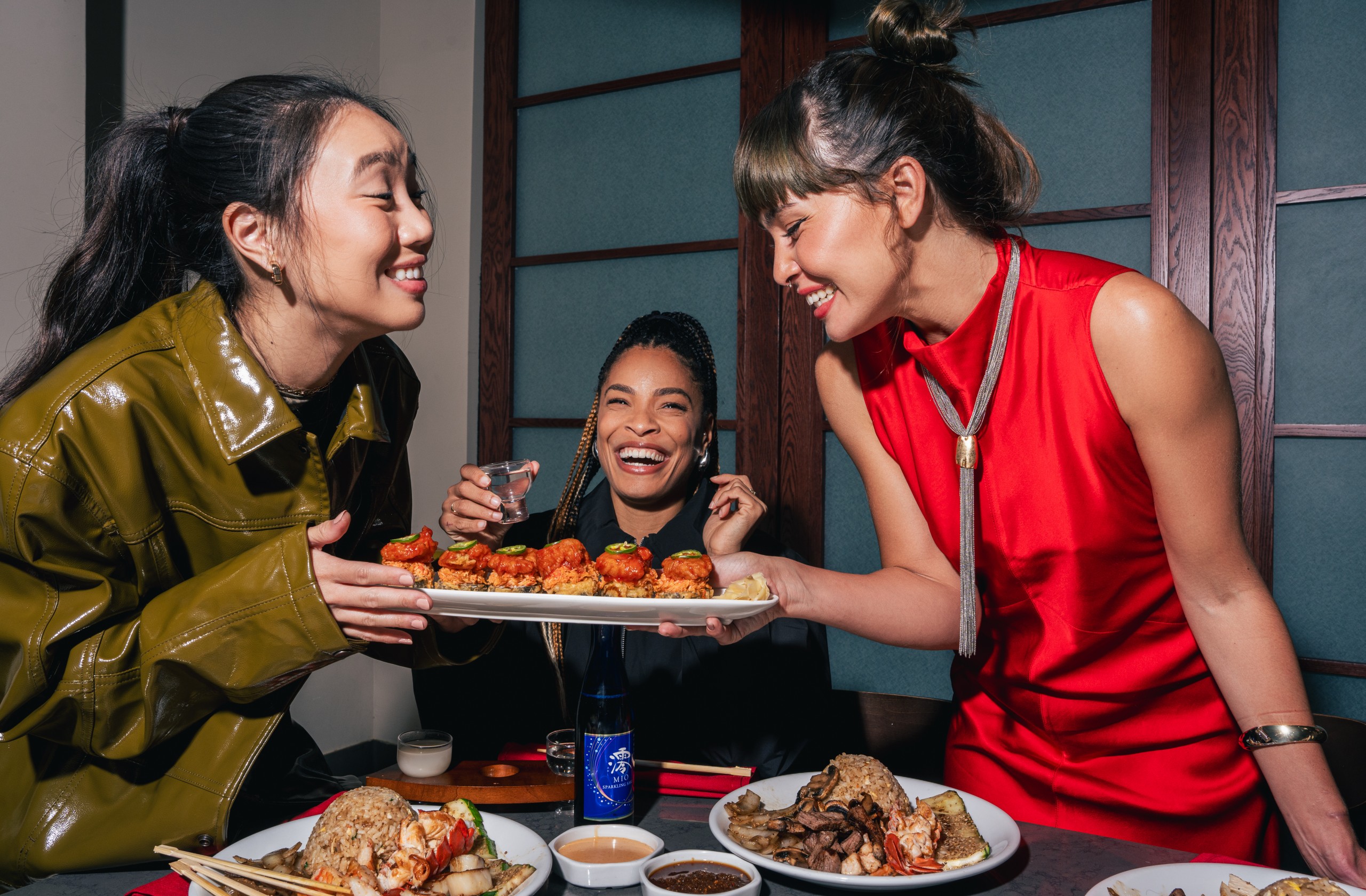 Three friends enjoying Benihana dishes and sake at a table. Plates of hibachi steak, hibachi chicken, grilled shrimp, fried rice, and vegetables. Dipping sauces are also visible on the table.